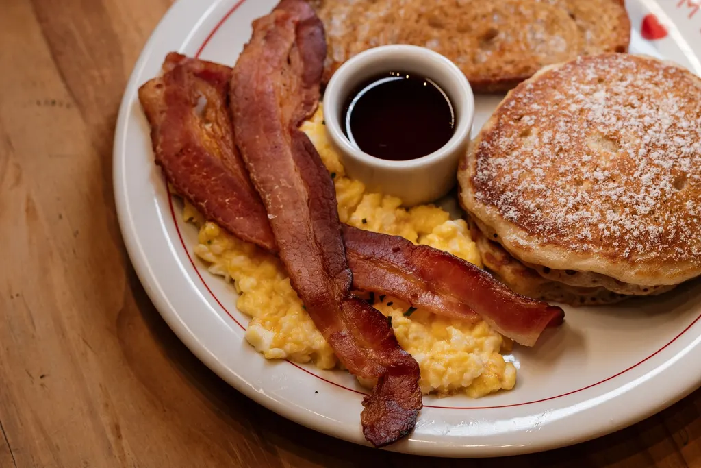 Breakfast plate of pancakes, bacon and scrambled eggs with a small cup of syrup on a wooden table