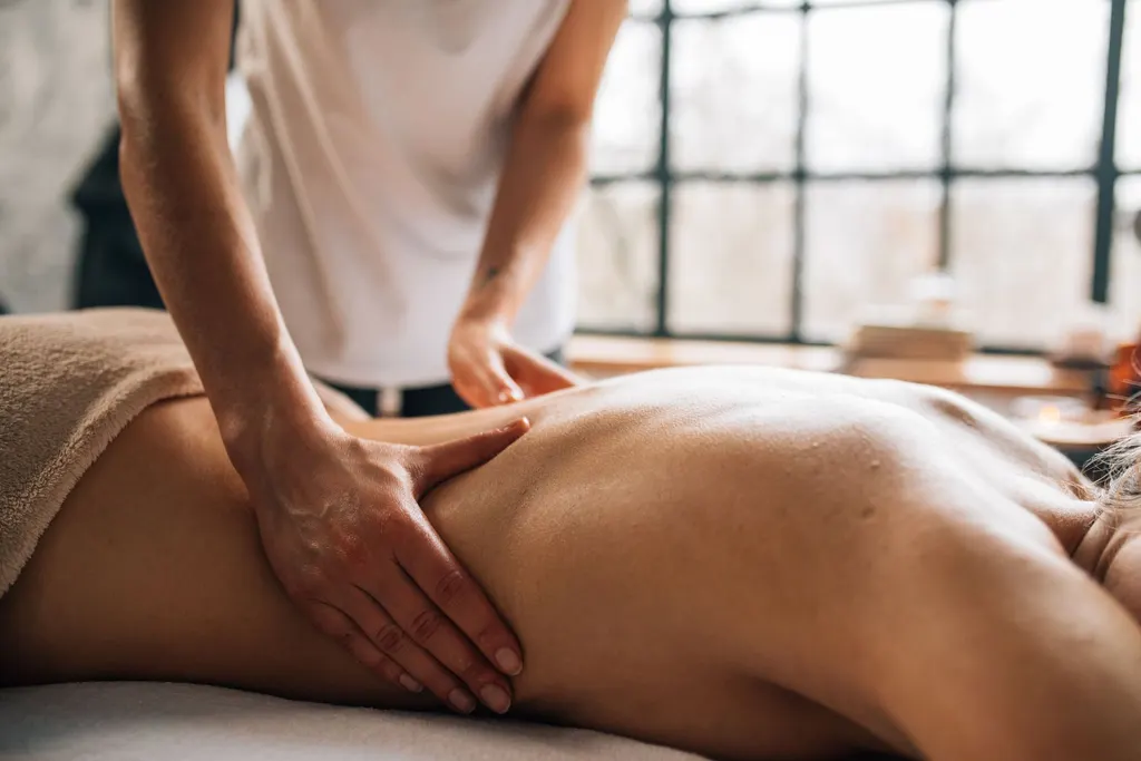 A massage therapist treating a client beside a sunlit window in a calm wellness studio.